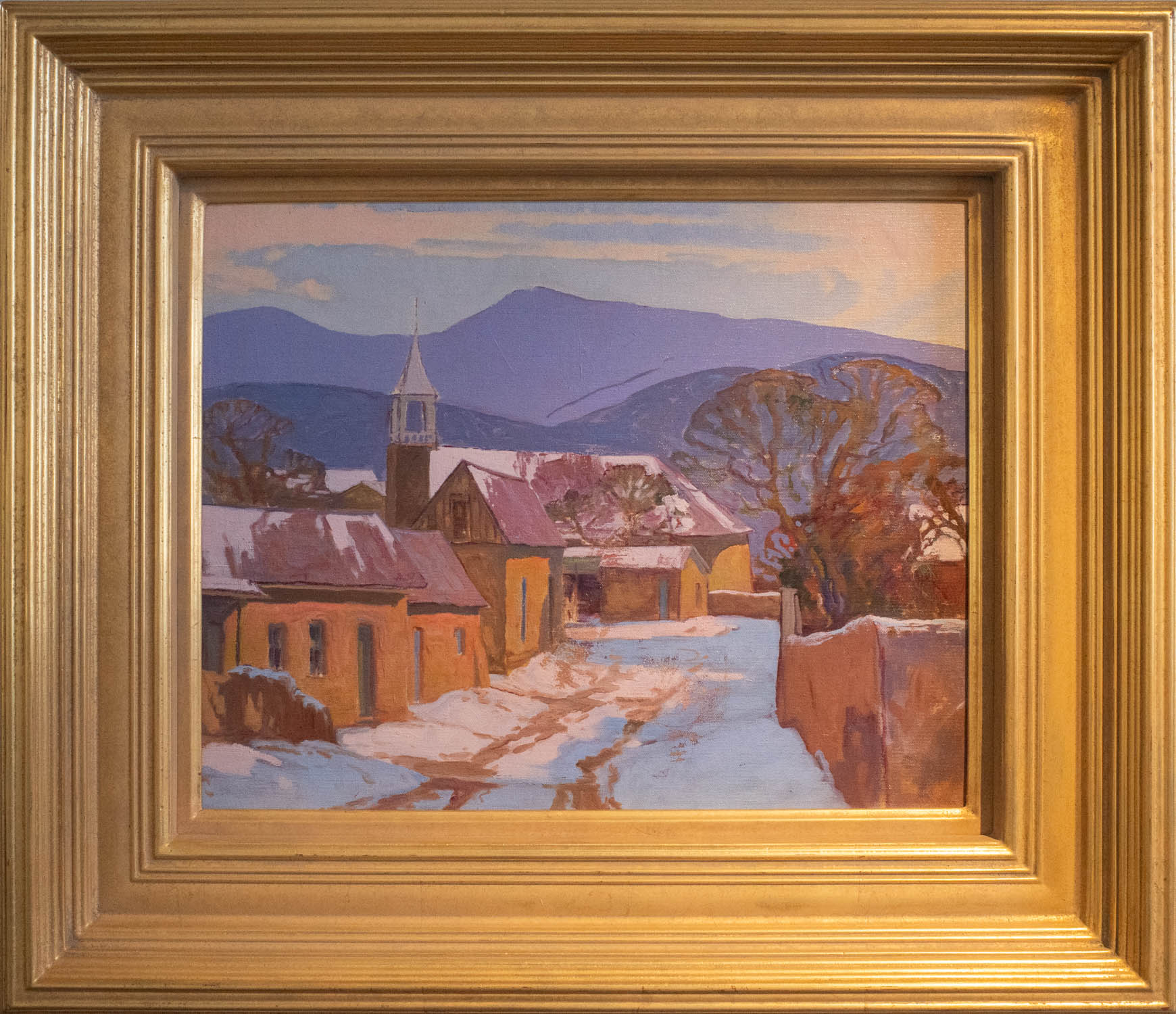 An impressionistic oil painting of a snowy village with a prominent church steeple, surrounded by snow-covered buildings against a backdrop of mountains under a purple and blue sky, presented in a detailed gold frame. Title: Pecos, New Mexico.